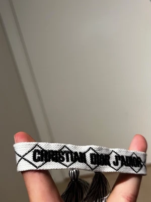 Christian Dior J'Adior armband - Snyggt vävt armband från Christian Dior med texten 'Christian Dior J'Adior' i svart på vit bakgrund. Armbandet har dekorativa tofsar i ändarna och ett coolt grafiskt mönster. Perfekt accessoar för att lyfta din outfit.