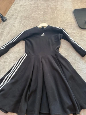 Svart Adidas klänning med ränder - Sportig svart klänning från Adidas med klassiska vita ränder längs ärmar och kjol. Klänningen har lång ärm, rund halsringning och ett utsvängt kjolparti. Adidas-logga på bröstet. Perfekt för en avslappnad och trendig look.
