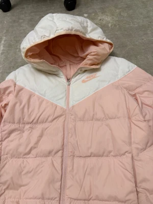 Nike rosa och vit pufferjacka - Säljer en snygg pufferjacka från Nike i ljusrosa och vitt. Jackan har huva, dragkedja framtill och ett broderat Nike-logotyp på bröstet. Tillverkad i ett quiltat material som håller värmen under kalla dagar. Perfekt för dig som vill ha en sportig och trendig vinterjacka.