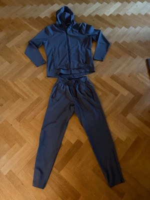 Grått träningsset från Under Armour - Säljer ett grått träningsset från Under Armour med matchande hoodie och byxor. Hoodien har dragkedja, huva och två fickor framtill. Byxorna har resår i midjan och smal passform. Perfekt för löpning eller gym. Jackan är M och byxorna S