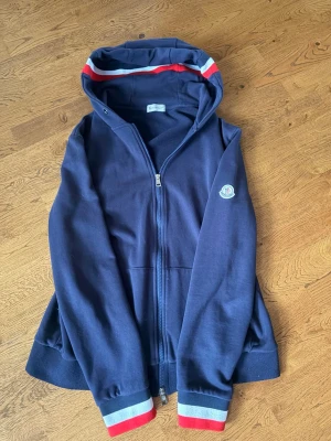 Moncler hoodie - Snygg marinblå moncler hoodie med dragkedja och huva. Röd och vit rand på huvan samt vid ärmsluten. Moncler-logga på ärmen. Perfekt för en avslappnad och trendig look! Pris går absolut att diskuteras och kan vara öppen för eventuella byten!!!