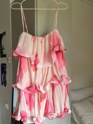 Kort volangklänning med rosa mönster - Säljer en superfin kort klänning med tunna axelband och volangdetaljer. Klänningen har ett plisserat tyg i vitt och rosa med stora blommiga inslag. Perfekt för sommardagar och har en luftig, loose passform. Har aldrig använt den då den inte passade, så den är helt ny och oanvänd✨