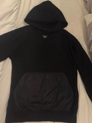 Svart hoodie med ficka och huva - Snygg svart hoodie med stor känguruficka framtill och klassisk huva. Hoodien har en enkel design med en liten logga på bröstet och är perfekt för en avslappnad stil. Materialet ser ut att vara en mix av bomull och syntet, vilket gör den både mjuk och slitstark. Pris kan diskuteras vid snabb affär 