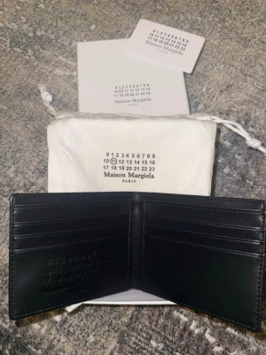 Svart läderplånbok Maison Margiela - Stilren svart plånbok i äkta läder från Maison Margiela. Plånboken har flera kortfack och präglad logotyp på insidan. Kommer med originalpåse och kort. Perfekt för dig som gillar minimalistisk design och hög kvalitet.