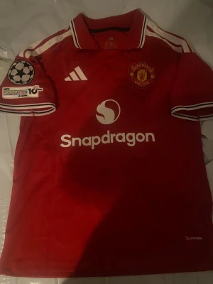 Manchester United matchtröja röd - Säljer en röd Manchester United matchtröja från Adidas med vita detaljer och klubbmärke på bröstet. Tröjan har Champions League-märke på ärmen, sponsorlogga på magen och spelarnamn B. Fernandes samt nummer 8 på ryggen. Perfekt för fotbollsfans!