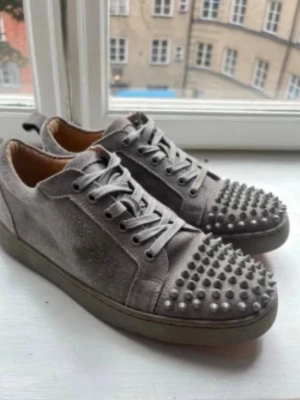 Gråa sneakers med nitar och röd sula - Unika grå sneakers i mocka med coola nitar på tån och klassisk röd sula. Skorna har grå snörning och en låg profil som ger en modern vibe. Perfekta för dig som vill sticka ut med detaljer och edgy stil