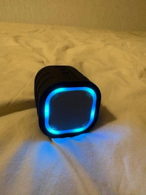 Svart bärbar högtalare med LED-ljus - Cool bärbar högtalare i svart med fyrkantig form och blå LED-belysning runt gallret. Smidig storlek som får plats i handen, perfekt för att ta med sig överallt. Tillverkad i plast och metall med tydliga knappar på ovansidan.