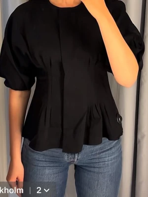 Svart blus med puffärm och markerad midja - Trendig svart blus med rund halsringning, puffärmar och markerad midja. Blusen har en dekorativ knappdetalj i sidan och lätt utsvängd nederkant. Perfekt att styla med jeans eller kjol för en snygg look.