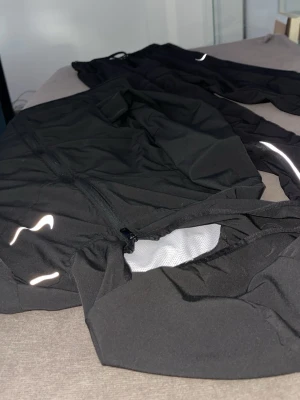 Svart träningsset från Nike - Säljer ett svart träningsset från Nike med jacka och byxor. Setet har reflekterande detaljer och dragkedja på jackan. Tillverkat i ett lätt och funktionellt material som passar perfekt för löpning eller annan träning. Snygg och sportig design med diskret Nike-logga.