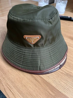 Olivgrön bucket hat  - Snygg olivgrön bucket hat från Prada i glansigt nylonmaterial med brunt läderkantband och den klassiska trekantiga Prada-loggan framtill. Perfekt accessoar för en trendig streetwear-look. ALDRIG ANVÄND