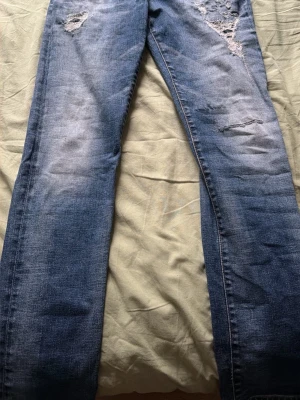 Blå slitna jeans  - Säljer ett par blå jeans med slitningar. Jeansen har en klassisk femficksmodell och raka ben. Materialet är denim i bomull och de har en snygg tvättad look som ger en avslappnad vibe. 30/30