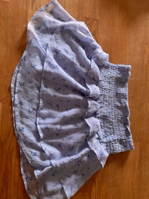 Blå volangkjol från Gina Tricot - Supersöt blå kjol från Gina Tricot med volanger och smock i midjan. Kjolen har ett blommigt mönster i blå toner och är gjord i ett lätt och luftigt material, perfekt för varma dagar. Passar perfekt till en enkel topp eller stickad tröja.