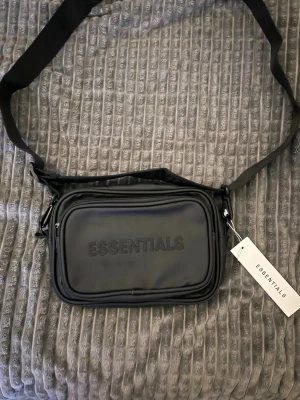 Svart axelväska från Essentials - Snygg svart axelväska från Essentials med justerbar rem och präglad logga på framsidan. Väskan har två fack med dragkedja och en stilren, rektangulär form. Tillverkad i ett slätt, tåligt material som passar perfekt till din streetwear-look.