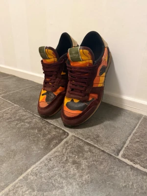 Färgglada rockrunners med camouflage mönster  - Unika rockrunner sneakers från Valentino med färgstarkt camomönster i orange, gul, svart och vinrött. Skorna har låga skaft, vinröda snören och detaljer i mocka och textil. Snygg kontrast mellan färgerna och cool design som sticker ut. Skicken på skorna är inte jättebra förutom om man kollar riktigt nära annars på fötterna är de perfekta skicka 7/10