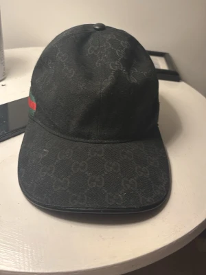 Svart keps med GG-mönster från Gucci - Snygg svart keps från Gucci med det klassiska GG-monogrammönstret över hela kepsen. På sidan finns en grön och röd rand som ger extra lyxig känsla. Kepsen har böjd skärm och justerbar rem bak. Perfekt accessoar för en trendig streetwear-look.