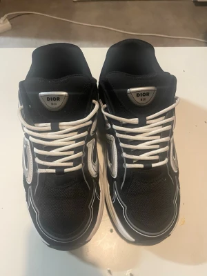 Di0r B3O svarta sneakers - Snygga Di0r B3O sneakers i svart med vita och grå detaljer. Skorna har en chunky vit sula, mesh och läderpaneler samt vita skosnören. Stora reflekterande Di0r-loggor på sidorna ger en modern vibe. Perfekta för dig som vill sticka ut med exklusiva sneakers.