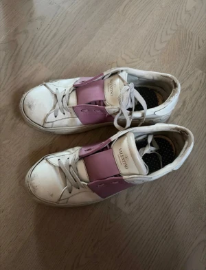 Valentino vita sneakers med rosa detalj - Snygga vita sneakers från Valentino med bred rosa läderdetalj över snörningen. Klassisk låg modell med vita skosnören och nitar på hälen. Använda men går att fixa hos skomakare. Har bytt ut sulorna och hittar ej dom originala tyvärr.