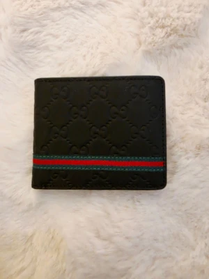Svart plånbok med GG-mönster - Snygg svart plånbok i läder med präglat GG-mönster och en klassisk grön-röd rand på framsidan. Plånboken har flera kortfack och ett myntfack på insidan. Perfekt för dig som vill ha en stilren och trendig accessoar.