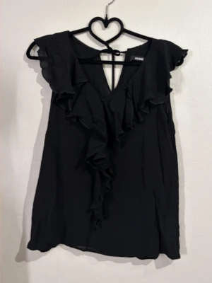 Svart blus  - Snygg svart blus med djup v-ringning och volangdetaljer framtill. Ärmlös modell med lätt transparent tyg och knyt i nacken. Perfekt för dig som gillar en feminin och trendig look.