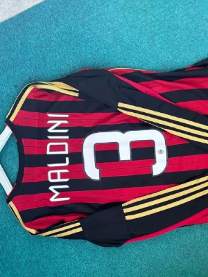 AC Milan matchtröja Maldini - Säljer en långärmad AC Milan matchtröja från Adidas med Maldini och nummer 3 på ryggen. Tröjan är röd och svart med guldfärgade ränder på ärmarna, klubbmärke och sponsorlogga på bröstet. Perfekt för dig som älskar fotboll och vill ha en ikonisk supportertröja.