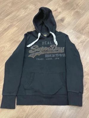 Svart hoodie från Superdry - Svart hoodie från Superdry med stor logga och japansk text på bröstet. Tröjan har huva med vita dragsnören och en klassisk känguruficka framtill. Mjuk insida och ribbade muddar vid ärmar och nederkant. Perfekt för en avslappnad streetwear-look.