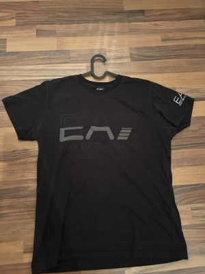 Svart EA7 Emporio Armani t-shirt - Svart t-shirt från EA7 Emporio Armani med stor logga på bröstet och mindre logga på ärmen. Klassisk passform med rund hals och korta ärmar. Perfekt för en stilren och modern look.