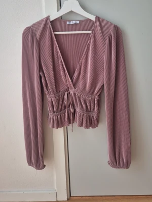 Rosa omlottopp från Zara - Säljer en ribbad rosa omlottopp från Zara med långa ärmar och knyt i midjan. Toppen har v-ringning och volangdetalj nedtill. Perfekt för dig som gillar en feminin och trendig stil.