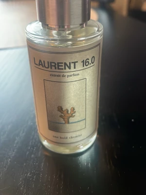 Laurent 16.0 Extrait de Parfum - En stilren parfymflaska med metallock och transparent glas. Etiketten är beige med svart text och en illustration av en korall. Doften heter Laurent 16.0 och är från One Bold Chemist. Perfekt för dig som gillar unika och moderna dofter. Doften är inspirerad av Luis Vuitton L’immensité! Använt Max 4ml. Vill få bort den snabbt därav pris
