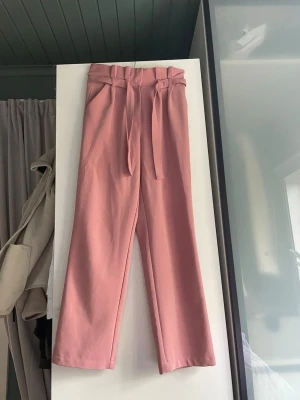 Vida rosa byxor med knytskärp - Säljer ett par vida byxor i en snygg rosa färg med hög midja och knytskärp i midjan. Byxorna har raka ben och rynkig linning för en trendig look. Perfekta för dig som vill ha något bekvämt men ändå stilrent.