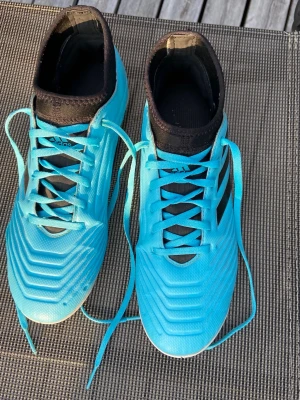 Blå fotbollsskor med högt skaft Adidas, stl 40 - Säljer ett par snygga fotbollsskor i klarblå färg med högt svart skaft och snörning. Skorna har en modern design med mönstrad ovansida och är tillverkade i syntetmaterial. Perfekta för dig som vill sticka ut på planen!