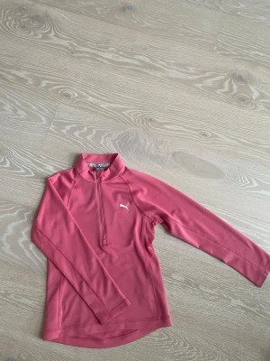 Rosa långärmad träningströja Puma - Rosa långärmad träningströja från Puma med halv dragkedja framtill och liten vit Puma-logga på bröstet. Tröjan har hög krage och är tillverkad i ett lätt, stretchigt material som passar perfekt för träning och sportaktiviteter.