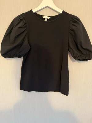 Svart topp med puffärmar från H&M i storlek S - Snygg svart topp med puffiga kortärmar och rund halsringning. Toppen har en enkel, stilren design och är perfekt för att lyfta vilken outfit som helst. Tillverkad i ett mjukt material som känns bekvämt mot huden.