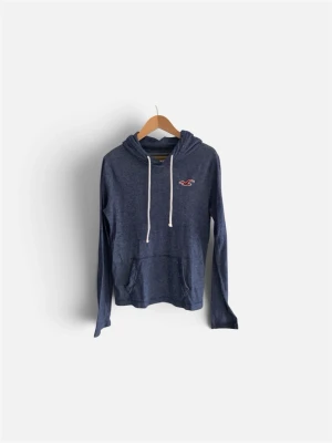 Blå hoodie med snörning från Hollister - Mysig marinblå hoodie från Hollister med vit snörning i huvan och klassisk röd logga på bröstet. Hoodien har känguruficka framtill och är perfekt för chill dagar. Skön och enkel stil som funkar till det mesta.