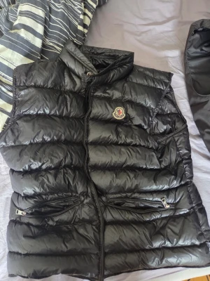 Svart dunväst från Moncler - Snygg svart dunväst från Moncler med glansig finish och klassisk logga på bröstet. Västen har hög krage, två dragkedjefickor framtill och quiltad design. Perfekt för lager-på-lager och streetwear-vibe.
