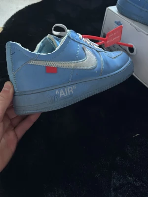 Nike x Off-White Air Force 1 blå - Säljer ett par Nike x Off-White Air Force 1 i blått läder med silverfärgad Swoosh, röda detaljer och den ikoniska röda taggen. Skorna har texten 'AIR' på sulan och tydliga Off-White-signaturer. Perfekta för dig som gillar streetwear och unika sneakers.