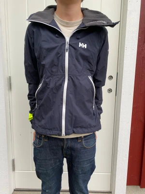 Mörkblå vindjacka från Helly Hansen - Snygg mörkblå vindjacka från Helly Hansen med vit dragkedja och logga på bröstet. Jackan har huva med gul insida, två fickor med dragkedja och justerbara ärmslut. Perfekt för blåsiga dagar och outdoor-aktiviteter.
