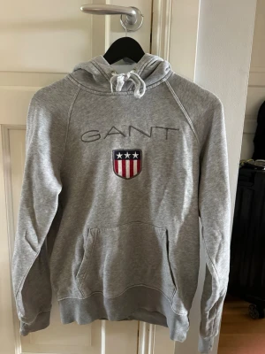 Grå hoodie från GANT med logga - Snygg grå hoodie från GANT med broderad logga och amerikanskinspirerat sköldmärke på bröstet. Klassisk känguruficka framtill, dragsko i huvan och ribbade muddar vid ärmar och nederkant. Perfekt för en avslappnad och sportig stil.