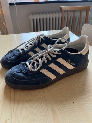Adidas Spezial mörkblå sneakers - Säljer ett par Adidas Spezial sneakers i mörkblå mocka med vita detaljer och klassiska tre ränder på sidan. Skorna har svart sula och vita skosnören. Guldtexten 'SPEZIAL' syns på sidan. Perfekta för dig som gillar retrostil och streetwear.