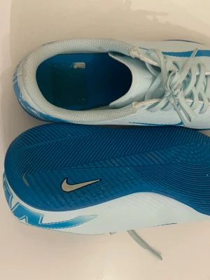 Nike blå-vita inomhusskor - Säljer ett par sportiga inomhusskor från Nike i ljusblått och vitt med blå sula och stora blå detaljer på hälen. Skorna har snörning och är tillverkade i syntetmaterial med meshfoder för bra ventilation. Perfekta för träning eller matcher inomhus.
