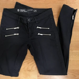 Svarta tight jeans med låg midja - Jeans med silver kedjor som detalj köpta i London. Tyvärr har dessa jeans fått en liten reva vid en utav fickorna på höger sida, men är annars felfria. Dragkedja vid fotknölen. I strl XS / 25.  