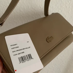 Ny LACOSTE plånbok/axelremsväska  - Mörk beige jättefin skinn plånbok/axelremsväska från Lacoste. Helt ny med lapp aldrig använt köpt i Usa för $248. Kan användas som plånbok utan straps som clutch eller som crossbody väska. Finns plats för kort och dragkedja med ficka. 