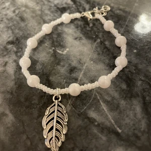 Armband - fjäder - Ett handgjort armband med pärlor och en fjäderberlock. Armbandet är justerbart och gjort av en elastisk tråd. Måttet är 19cm.
