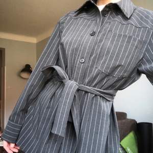 Snygg skjorta från weekday med en kostymkänsla! De tillkommer ett tyg skärp till som man kan knyta runt! Färgen är mörkgrå med vita stripes!