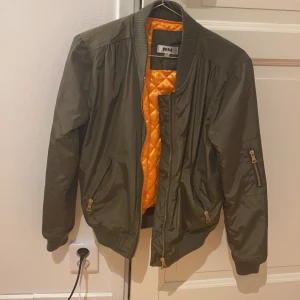 Bomberjacka  - Grön bomberjacka med gulddetaljer och orange insida. Nyskick! Hämtas upp i Bergsåker 