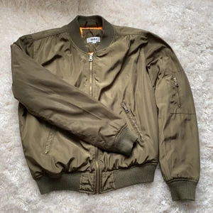 Grön bomber jacket - Bomber jacked med orange fodring. Jackan har en innerficka samt två praktiska ytterfickor