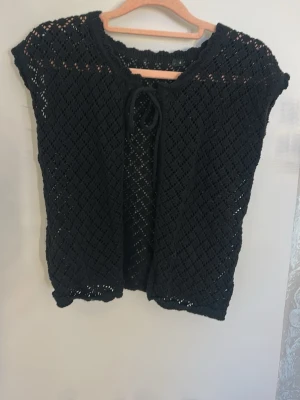 Svart virkad kofta med knytband - Snygg svart virkad kofta med korta ärmar och knytband framtill. Koftan har ett genombrutet mönster som ger en luftig känsla och passar perfekt över ett linne eller t-shirt. Materialet känns mjukt och följsamt.