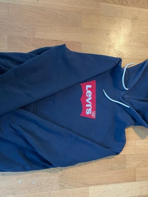 Mörkblå Levi's hoodie med logga - Snygg mörkblå hoodie från Levi's med den klassiska röda loggan på bröstet. Den är en M men passar S också bra .Hoodien har vit dragsko i huvan och en stor magficka framtill. Perfekt för en avslappnad och trendig stil. Tillverkad i mjukt bomullsmaterial. 