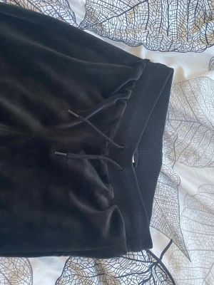 Svarta utsvängda mjukisbyxor - Säljer ett par svarta mjukisbyxor i mjukt velourmaterial med utsvängda ben och bred resår i midjan. Byxorna har dragsko i midjan för justerbar passform och en skön, avslappnad look. Perfekta för chill dagar eller när du vill vara extra bekväm.