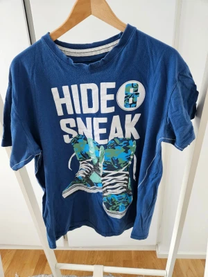 BLWR Blå t-shirt med tryck 'HIDE SNEAK' - Blå t-shirt med stort vitt tryck 'HIDE SNEAK' och motiv av sneakers i blått, turkost och zebra-mönster framtill. Rund halsringning och korta ärmar. Skön och avslappnad passform, perfekt för en chill stil.