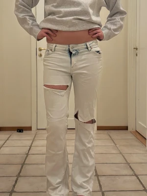 Vita ripped jeans med raka ben - Säljer ett par vita jeans med raka ben och stora slitningar över båda knäna. Jeansen har klassisk femficksmodell och knappgylf. Perfekta för en avslappnad och trendig look.